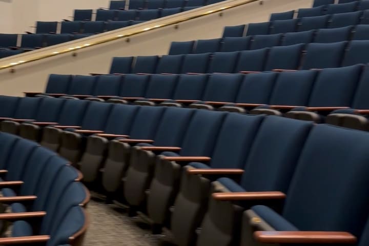 Curved rows of blue upholstered auditorium seats with wooden arms, climbing away from the stage.