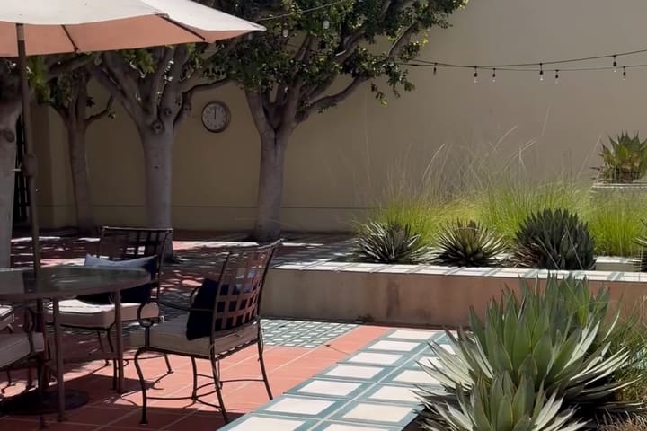 A courtyard with patterned tilework, agaves in raised beds, cafe tables under a parasol and string lights between trees.